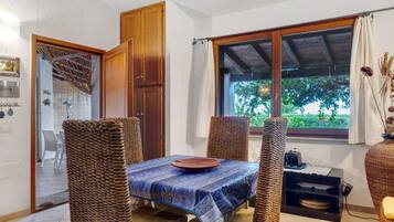 A dining area with a table set for two, surrounded by wicker chairs, a wooden door, and a window with a view of greenery.