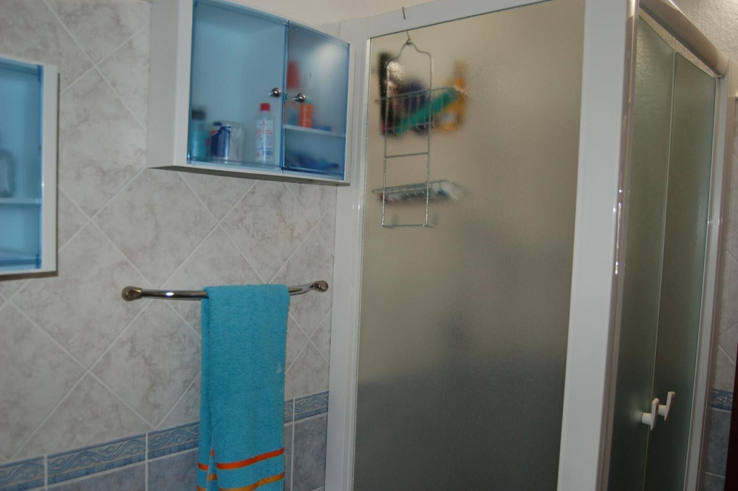A bathroom with a glass shower door, a towel rack with a blue towel, and a shelf with toiletries.
