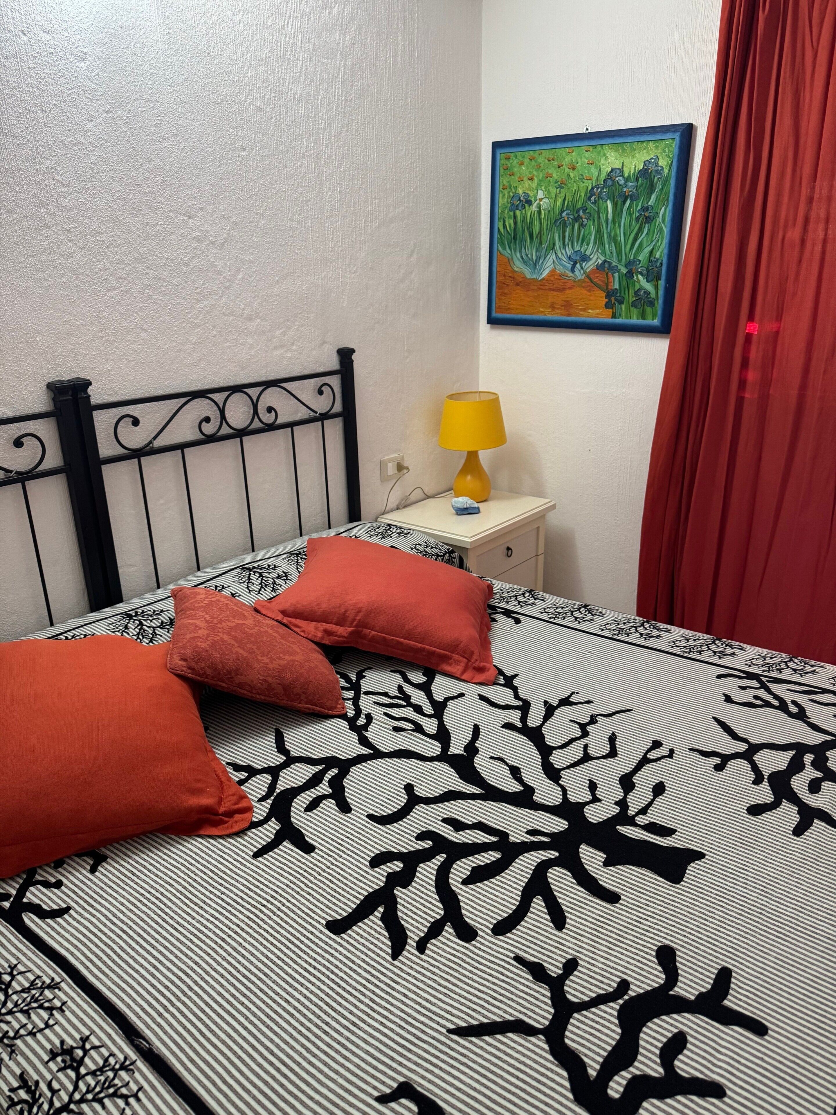 A bed with a striped bedspread and two red pillows, a nightstand with a yellow lamp, a painting on the wall, and a red curtain.
