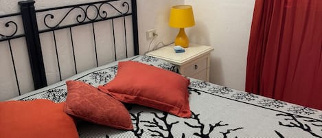 A bed with a striped bedspread and two red pillows, a nightstand with a yellow lamp, a painting on the wall, and a red curtain.
