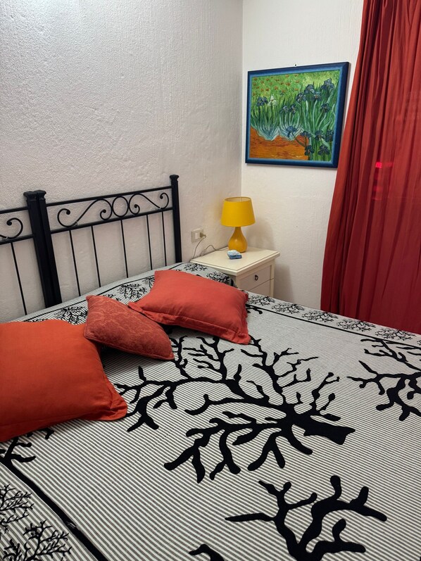 A bed with a striped bedspread and two red pillows, a nightstand with a yellow lamp, a painting on the wall, and a red curtain.
