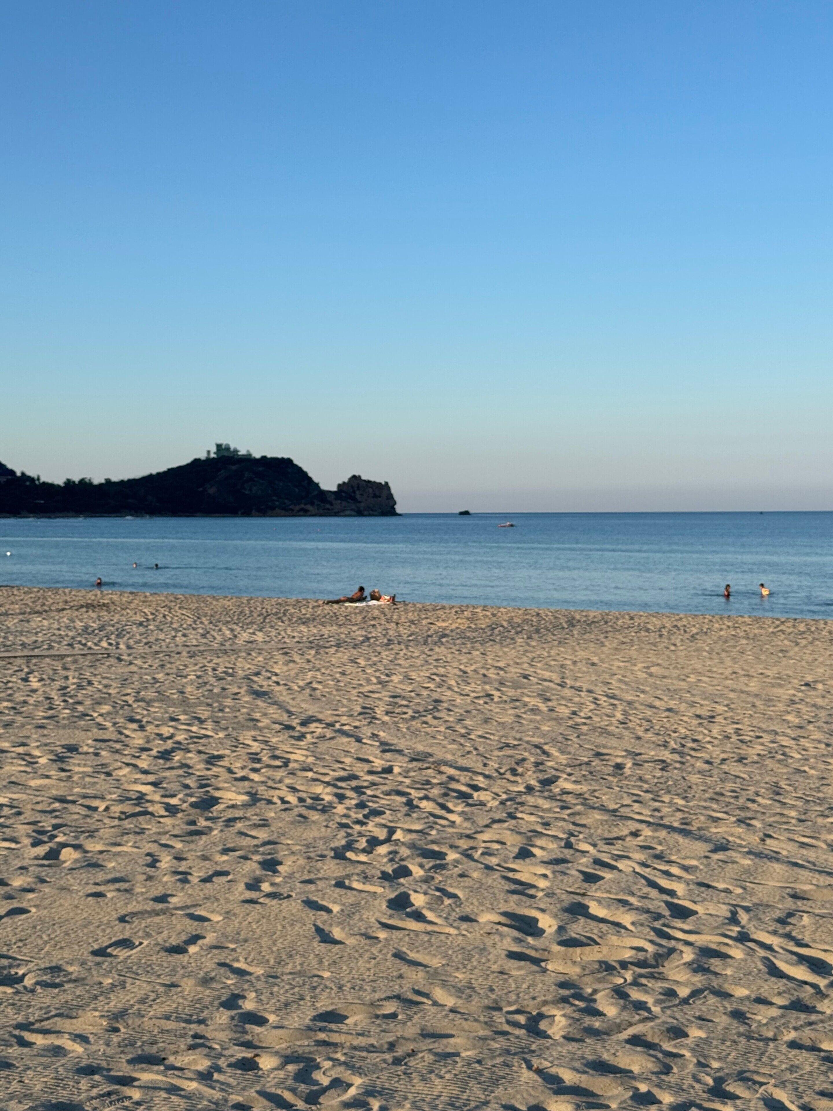 A sandy beach with footprints, a calm sea, and a distant rocky outcrop.