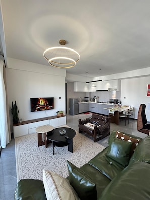 A modern living room with a flat-screen TV, leather sofa, and a dining area with a kitchen in the background. - Traveler's Loft- Park views near JKIA&SGR with Pool & Gym (Nairobi)