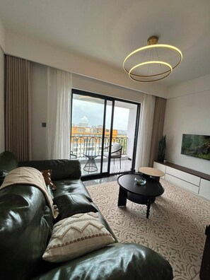 A modern living room with a leather sofa, a round coffee table, and a large window with a view of buildings. - Traveler's Loft- Park views near JKIA&SGR with Pool & Gym (Nairobi)