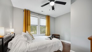 Une chambre à coucher comprenant un lit, une table de chevet, une lampe, un ventilateur de plafond et une fenêtre avec des rideaux.