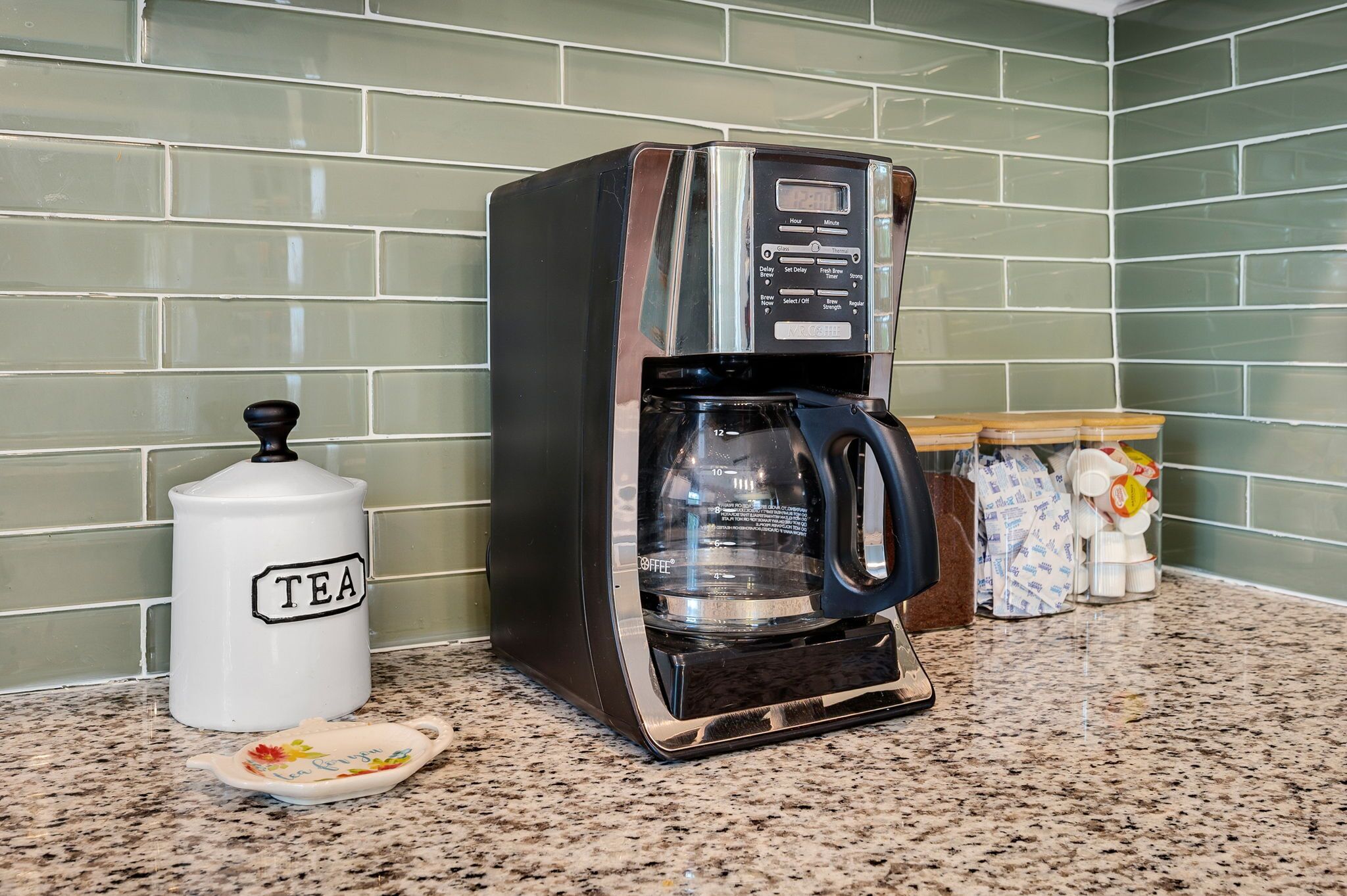A coffee machine with a digital display, a white mug labeled 'TEA', and a ceramic plate with a floral design.