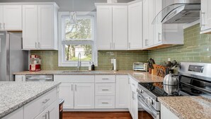 Une cuisine moderne avec des meubles blancs, un plan de travail en granit, une cuisinière en acier inoxydable et une fenêtre équipée de stores.