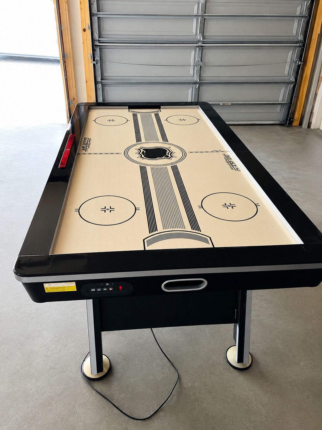 A pool table with a black frame and a beige playing surface, located indoors with a garage door in the background.