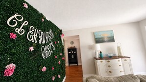 Ein Zimmer mit einer grunen Wand, die mit Pflanzen und Blumen bewachsen ist, eine weiße Kommode mit Schubladen, eine Lampe und ein Bild an der Wand.