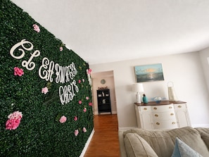 A room with a green wall covered in foliage and flowers, a white dresser with drawers, a lamp, and a painting on the wall. - Clearwater Oasis | Private Pool | 5mi from Beach (Pinellas County)