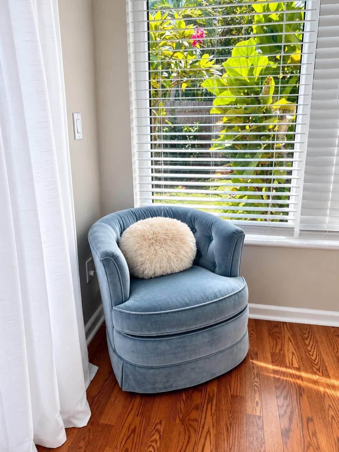 Ein Eckzimmer mit blauem Sessel, einem flauschigen Kissen, einem Fenster mit Jalousien und Holzdielen.