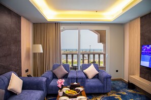 A hotel room with a balcony, two blue sofas, a dining table with fruit, and a TV on the wall.