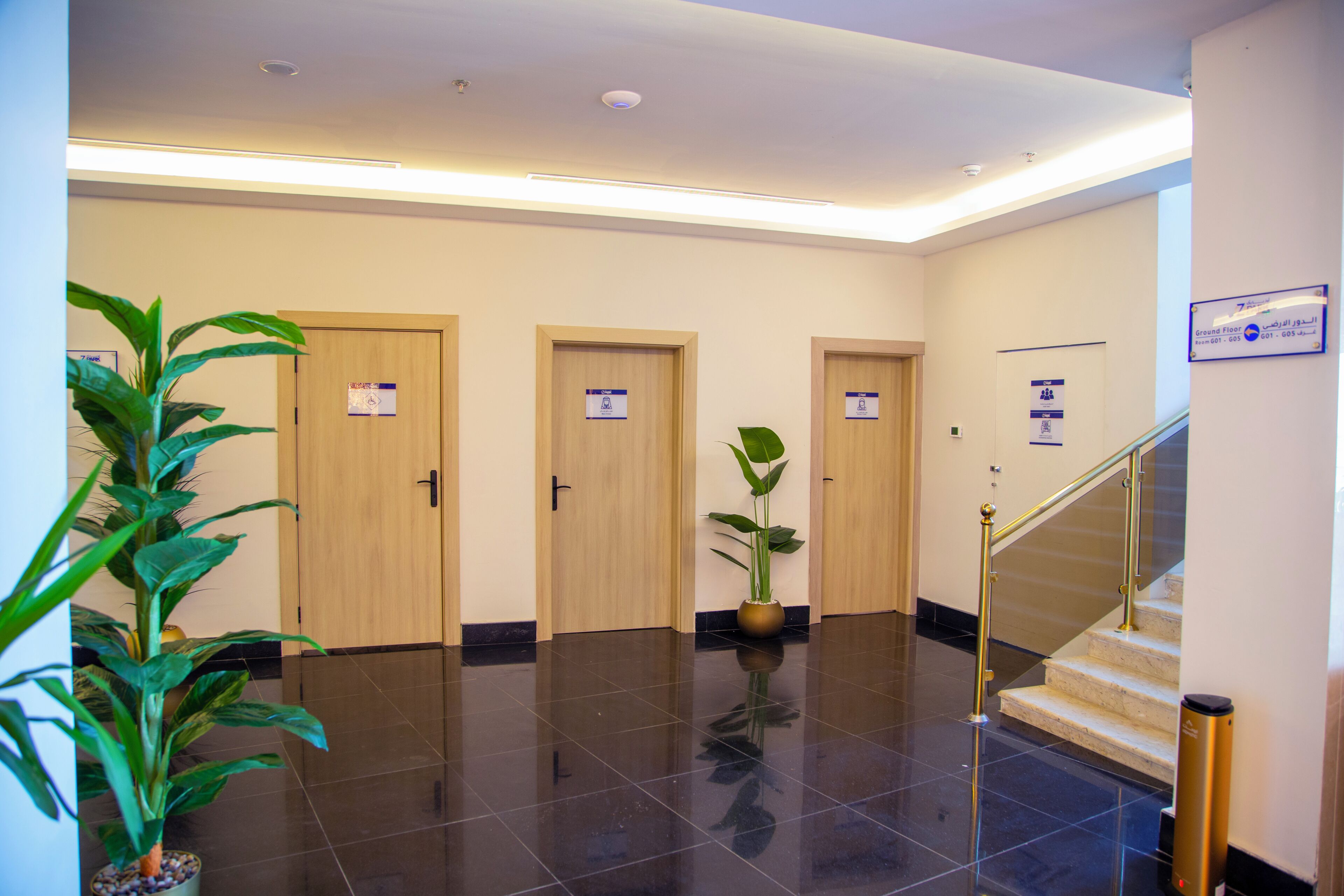 Un vestíbulo de hotel con piso pulido, tres puertas de madera, una escalera y plantas en macetas.