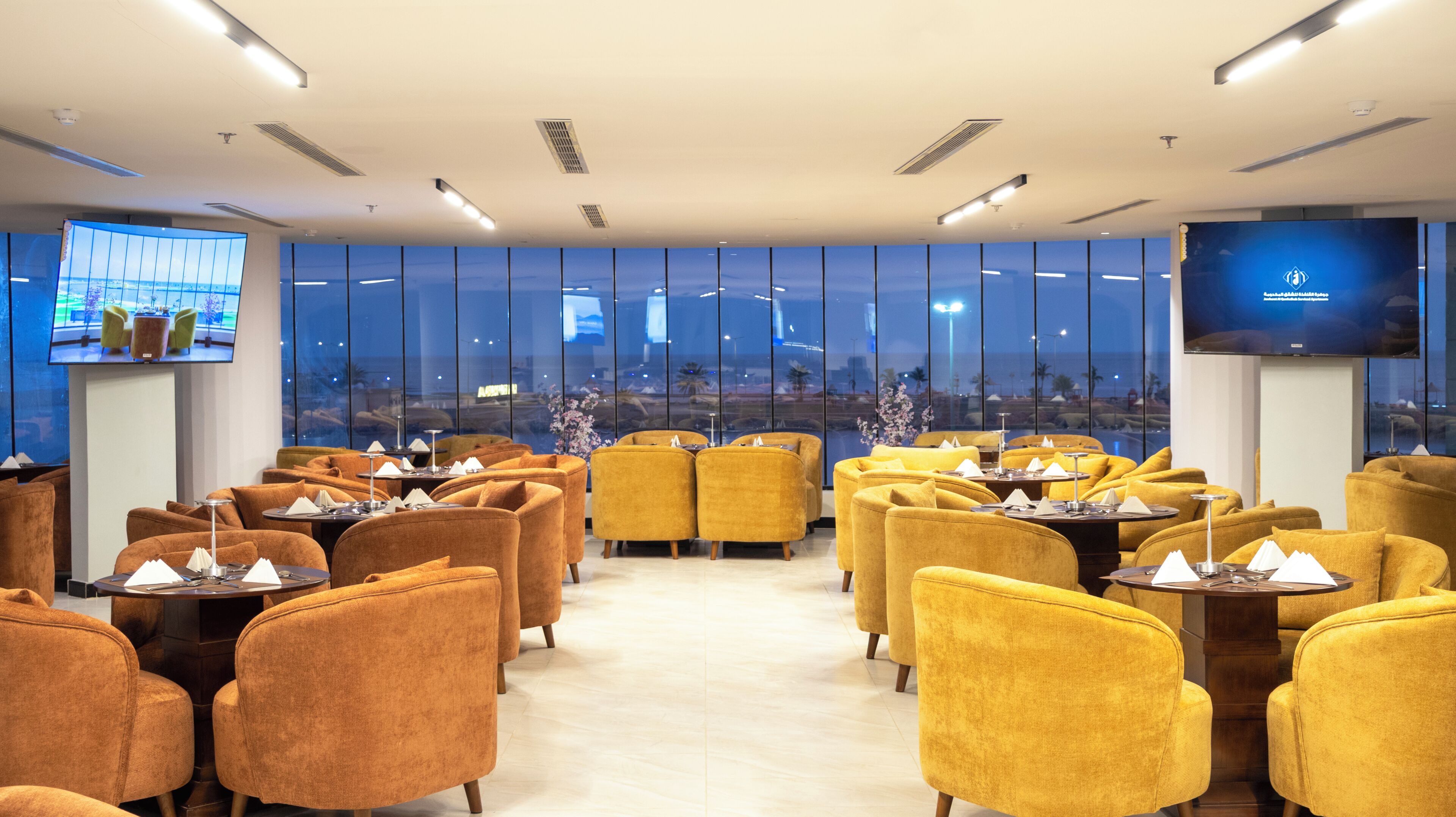 A hotel room with a large window, a flat-screen TV, and a dining area with yellow chairs and tables.
