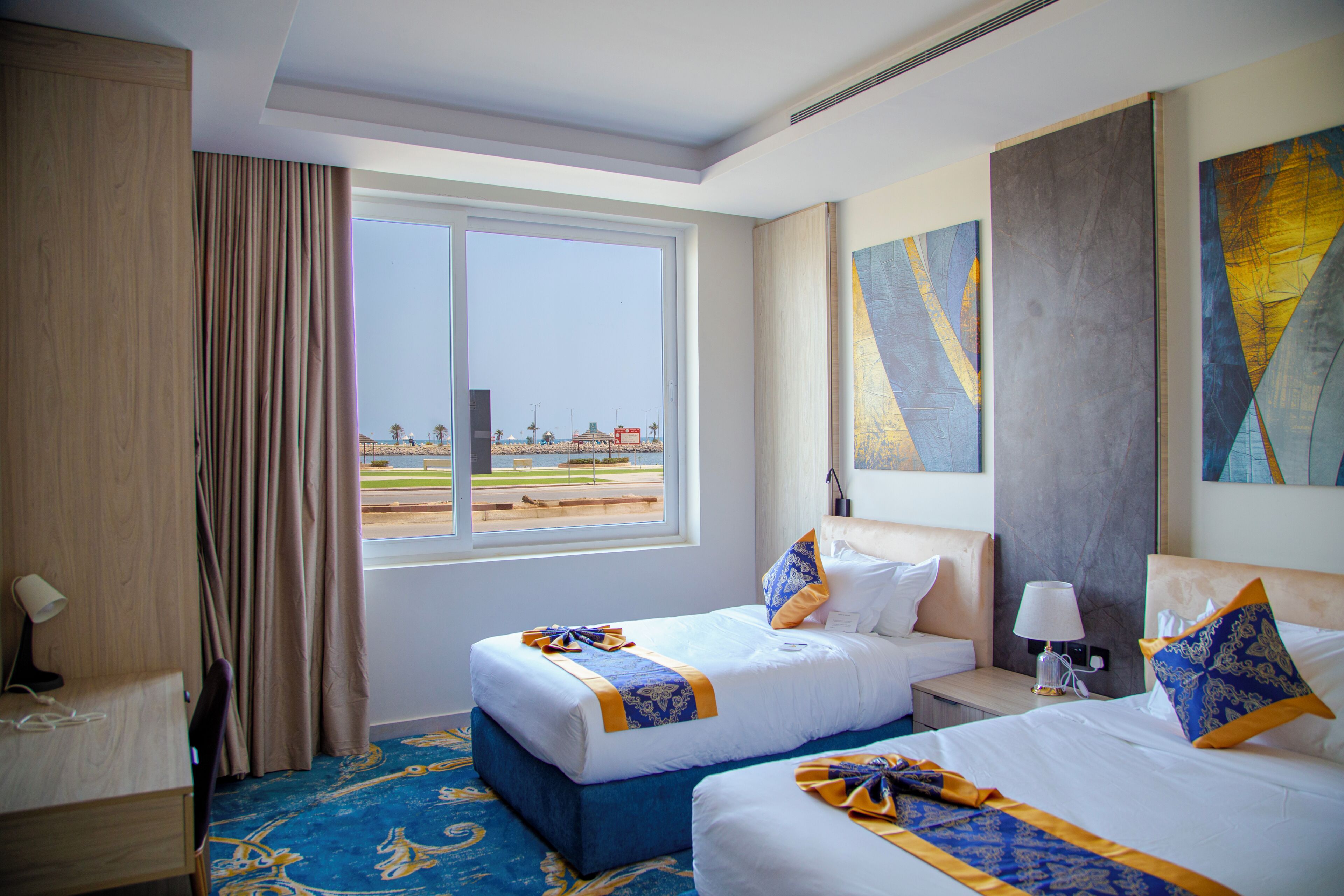 Quarto de hotel com duas camas, uma janela grande com vista para uma praia e um quadro na parede.