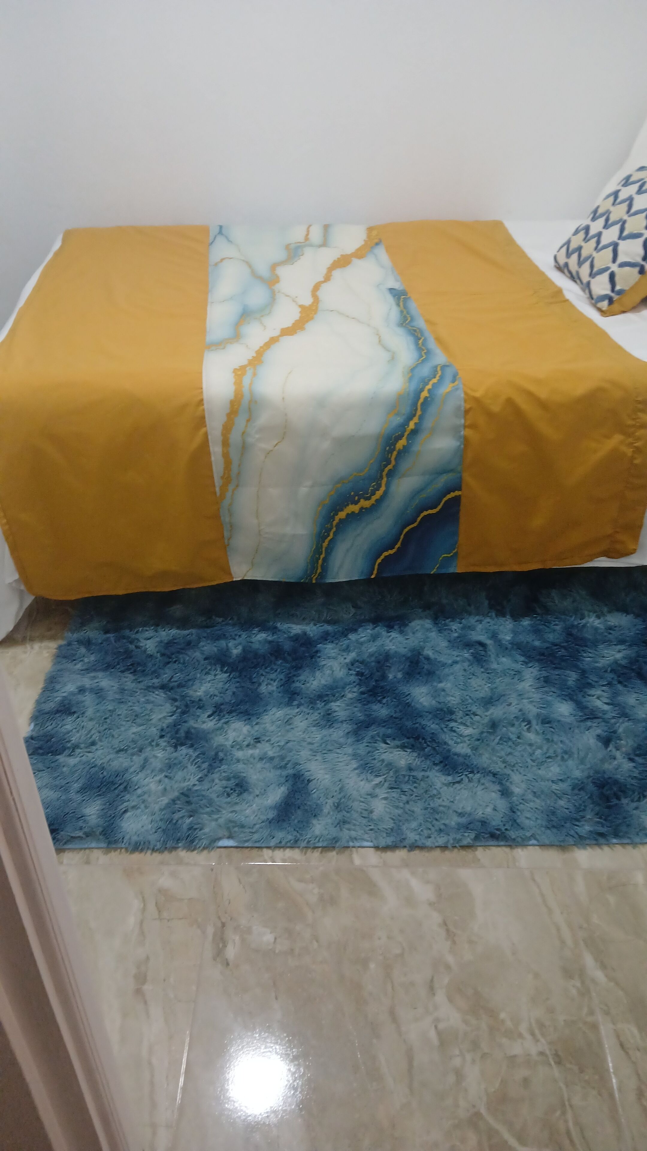 A bed with a yellow and white patterned bedspread, a blue fluffy rug, and a marble floor.