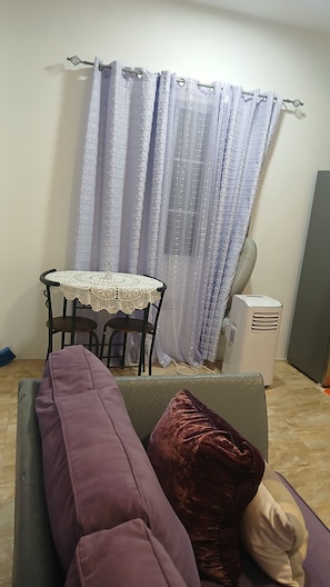 A room with a purple sofa, a small round table with a lace tablecloth, and a white air cooler.
