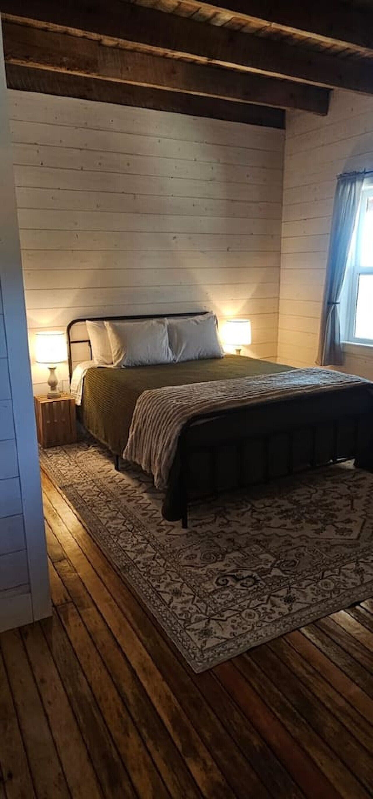 Un dormitorio con una cama, dos lámparas de noche, un tapete estampado, suelo de madera y una ventana con cortinas.