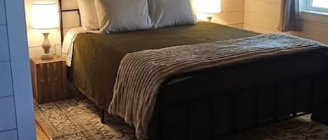 Un dormitorio con una cama, dos lámparas de noche, un tapete estampado, suelo de madera y una ventana con cortinas.