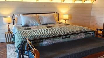 Una cama con estructura de madera, colcha y almohadas, una mesita de noche con lámpara y una alfombra estampada sobre piso de madera.