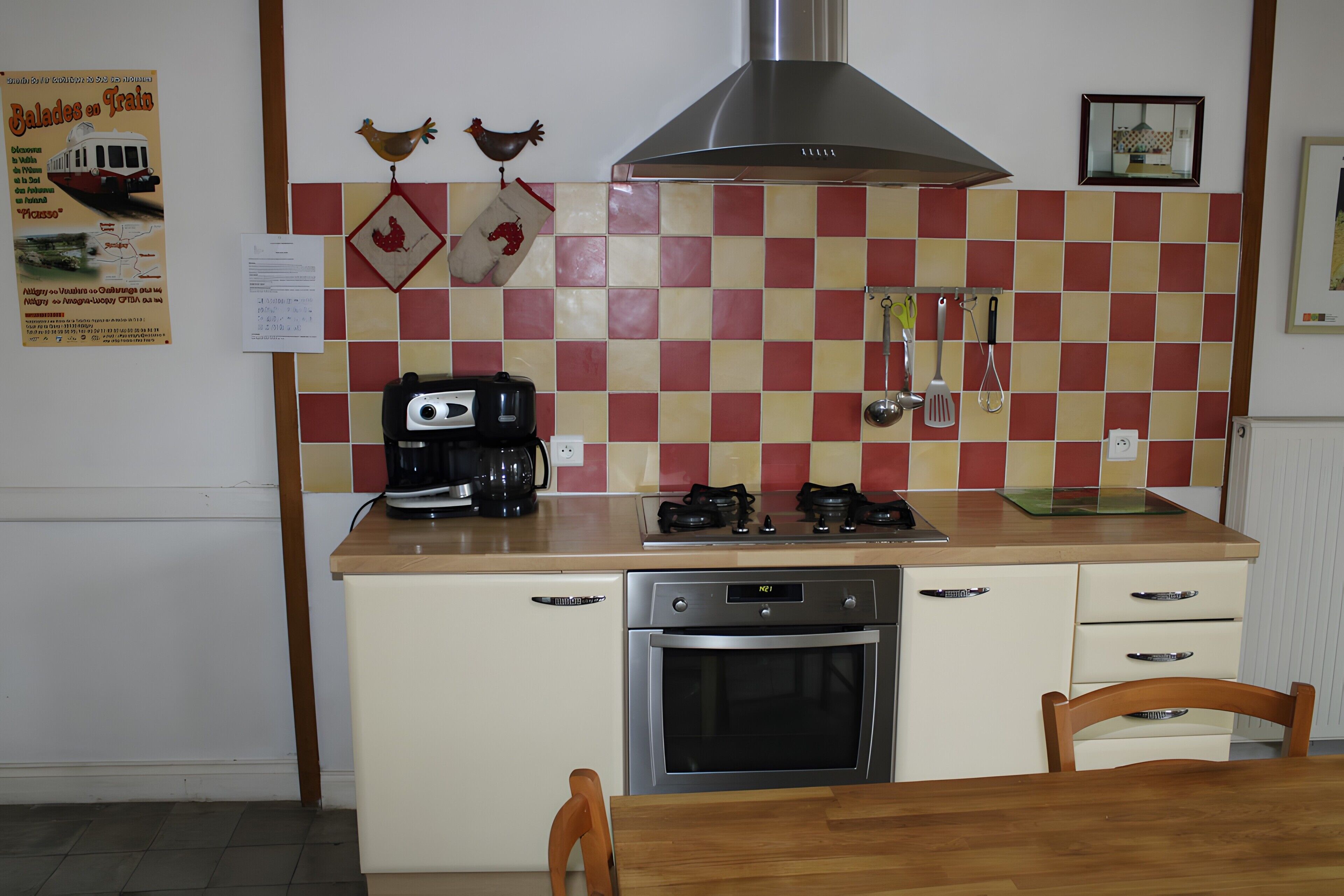 Een keuken met een fornuis, oven en koffiezetapparaat. Er zijn twee stoelen en een tafel.