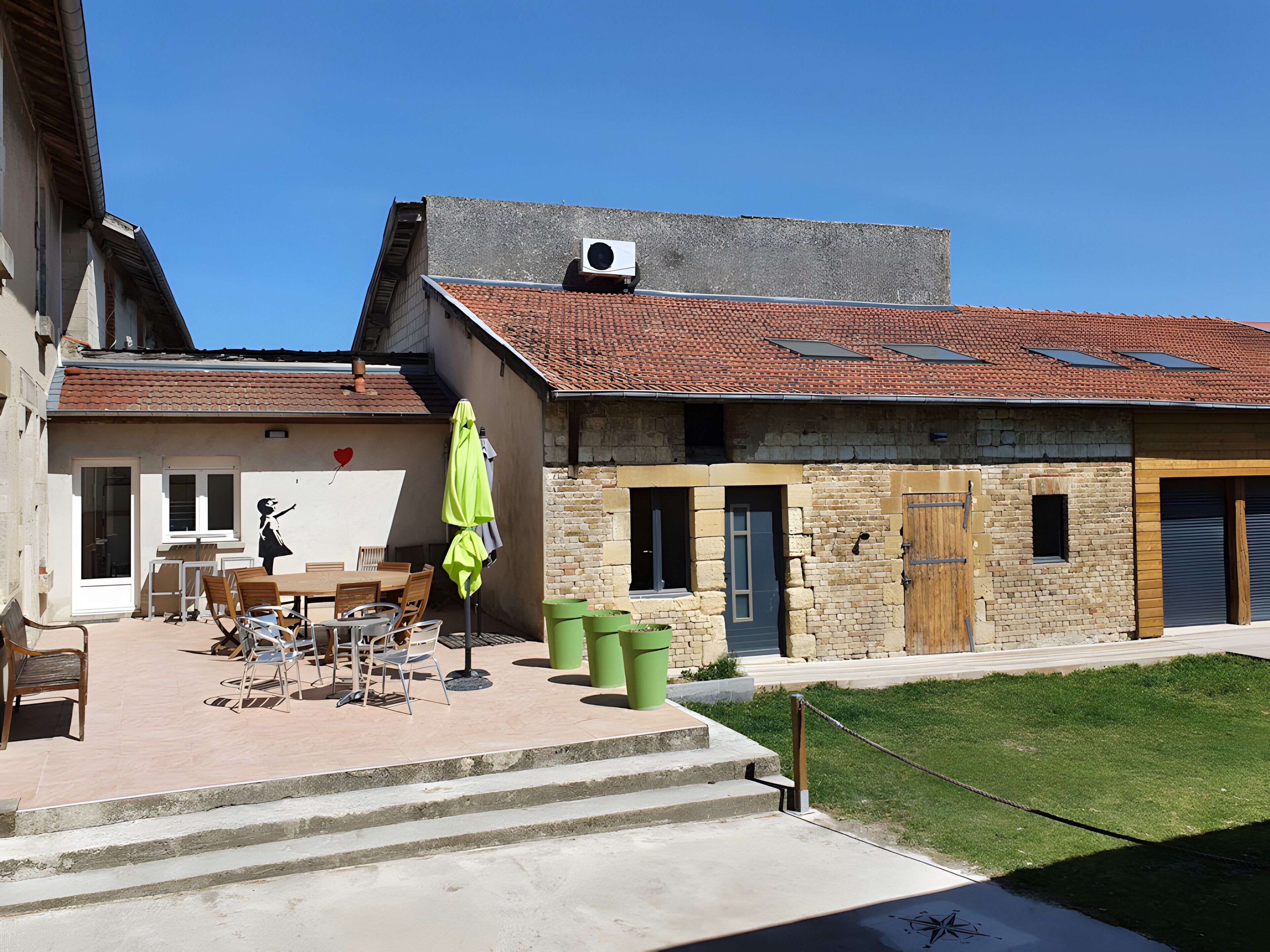 A building with a tiled roof, a covered outdoor seating area with chairs and tables, and a green trash bin.
