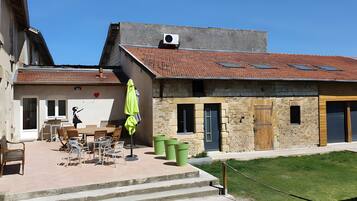 A building with a tiled roof, a covered outdoor seating area with chairs and tables, and a green trash bin.
