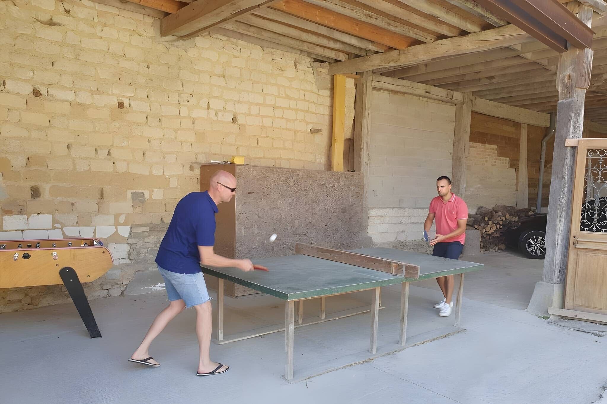 Twee personen spelen tafeltennis buiten, met een bakstenen muur en een houten constructie op de achtergrond.