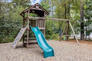 A wooden play structure with a slide and swings, surrounded by trees. - Spotted Feather: Escape and enjoy Hot Tub, Playset, Foosball, and Fireplaces. (Broken Bow)