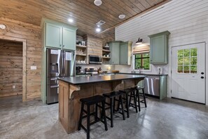 A kitchen with wooden walls, a central island with stools, stainless steel appliances, and a window with a view. - Spotted Feather: Escape and enjoy Hot Tub, Playset, Foosball, and Fireplaces. (Broken Bow)
