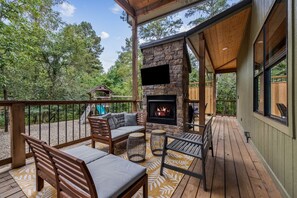 A covered deck with a stone fireplace, outdoor seating, and a playground in the background. - Spotted Feather: Escape and enjoy Hot Tub, Playset, Foosball, and Fireplaces. (Broken Bow)