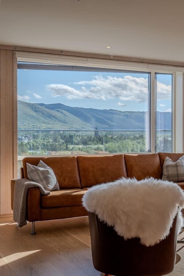 A modern living room with a brown leather sofa, a white furry ottoman, and a large window offering a view of a mountainous landscape.