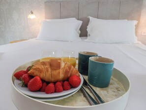 Ein Frühstücksbuffet mit Croissants, Erdbeeren, Himbeeren und Saft auf einem Tablett, im Hintergrund ein Bett mit Kissen.