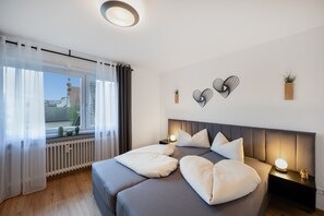 A modern bedroom with a large bed, bedside lamps, a view of the city through the window, and decorative wall art. - Apartment 'Weitblick' with Mountain View, Balcony, and Wi-Fi (Reute)