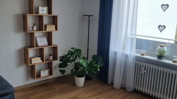 A room with a wooden floor, a bookshelf, a potted plant, a radiator, and a window with curtains. - Apartment 'Weitblick' with Mountain View, Balcony, and Wi-Fi (Reute)