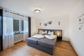 Quarto com cama, luminárias de cabeceira, radiador e janela com cortinas.
