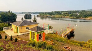 Een vakantiehuis met een terras dat uitkijkt over een meer, een steiger met een boot, en een beboste heuvel op de achtergrond.