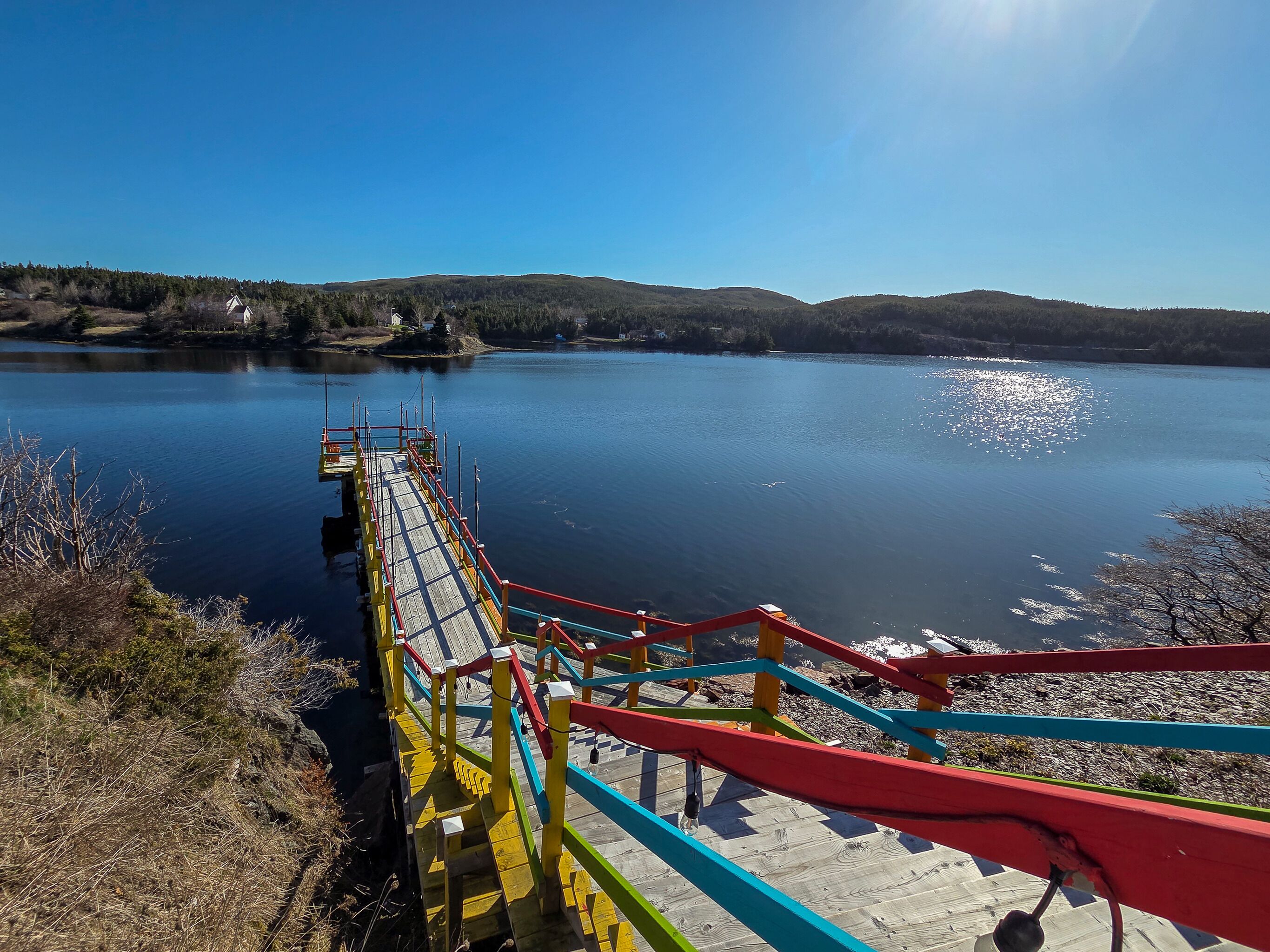 Un pont en bois, agrémenté de garde-corps colorés, mène à un lac paisible, entouré de collines.