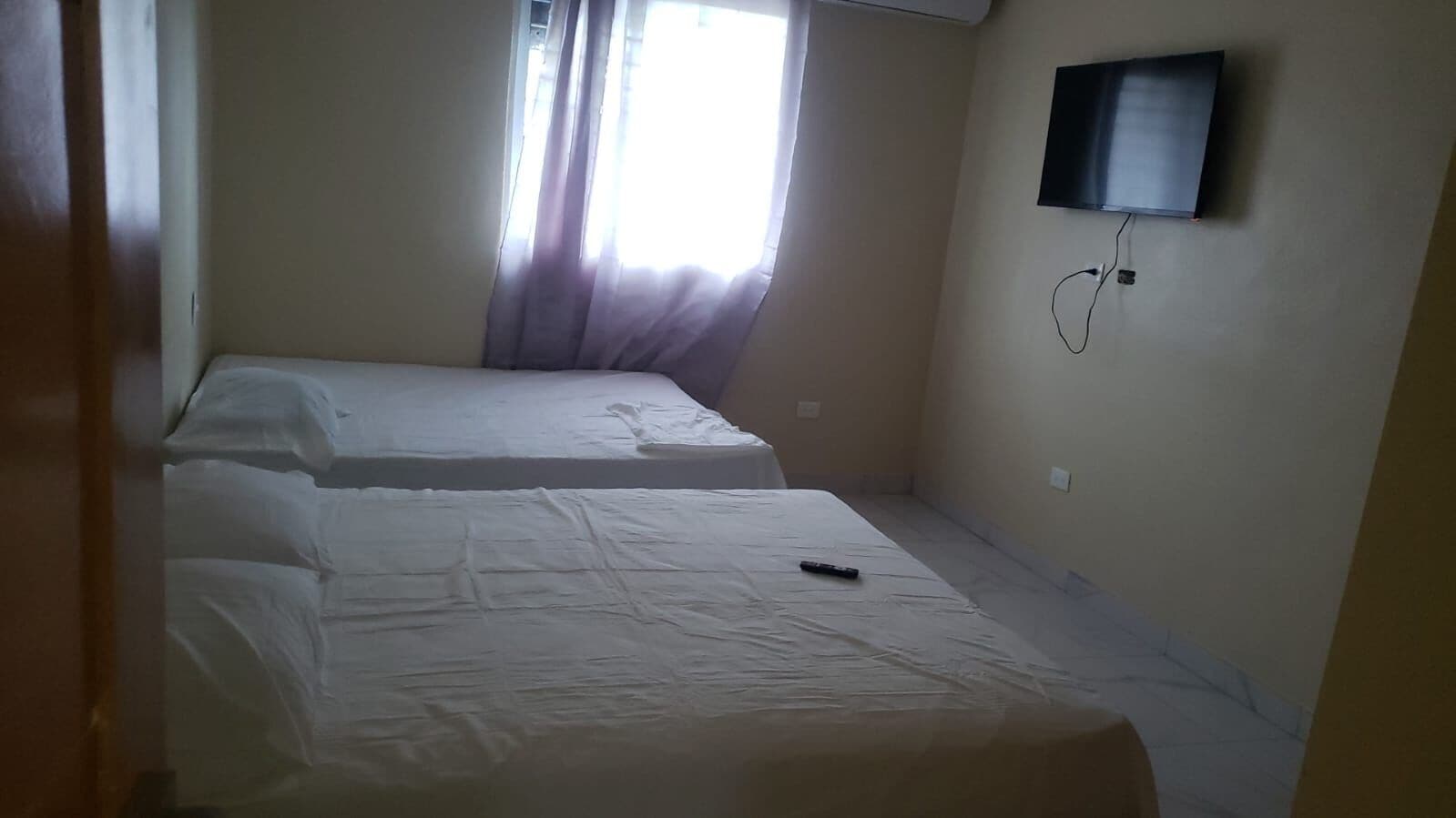 A room with two beds, a television mounted on the wall, and a window with curtains.