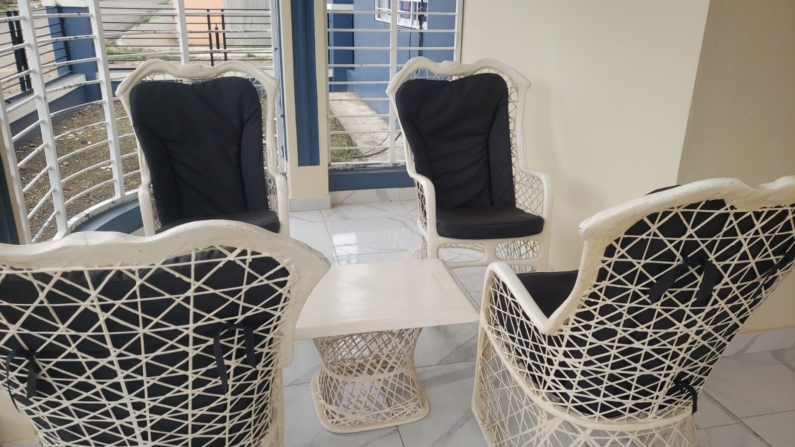 A balcony with white wicker chairs and a small table, overlooking a blue railing and a building.