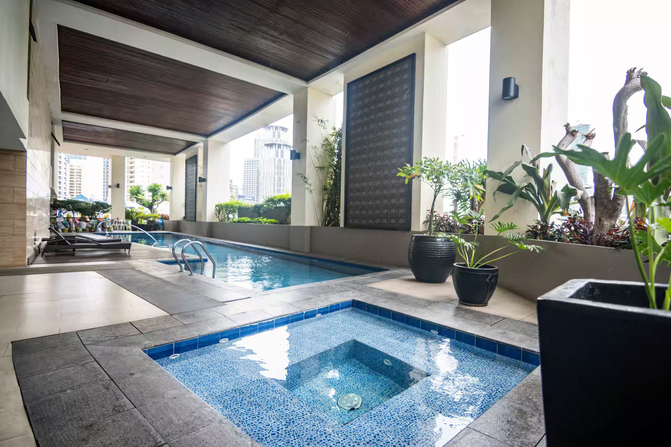 An indoor swimming pool with a tiled floor, surrounded by potted plants and a view of tall buildings.