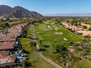 A golf course with a winding path, surrounded by residential buildings with red-tiled roofs, and mountains in the background. - Fairway Peaks at PGA West | Mountain & Golf Views (La Quinta)
