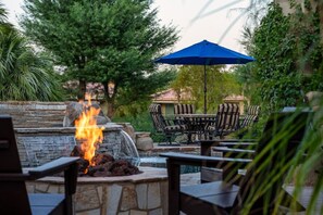 A patio area with a stone fire feature, outdoor furniture, and a blue umbrella. - Fairway Peaks at PGA West | Mountain & Golf Views (La Quinta)