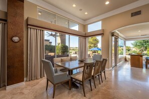 A spacious dining area with a large table and chairs, a clock on the wall, and large windows offering a view of the outdoors. - Fairway Peaks at PGA West | Mountain & Golf Views (La Quinta)