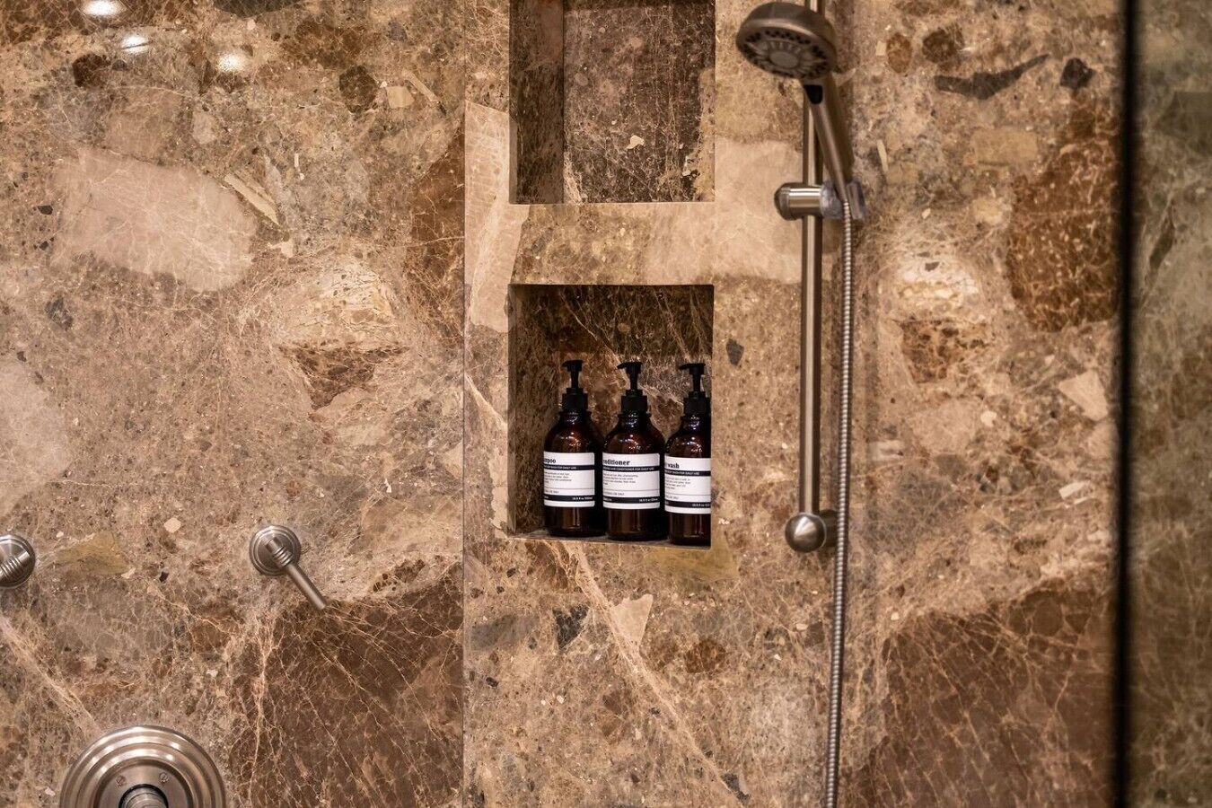 A bathroom with a marble wall, a showerhead, and a shelf with three bottles of liquid.