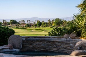A pool with a stone wall, surrounded by a golf course with palm trees and mountains in the background. - Fairway Peaks at PGA West | Mountain & Golf Views (La Quinta)