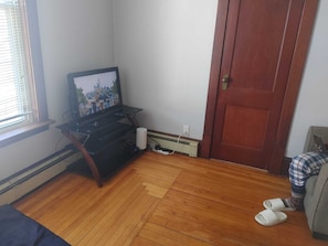 A room with a wooden floor, a television on a stand, a door, and a window with blinds.