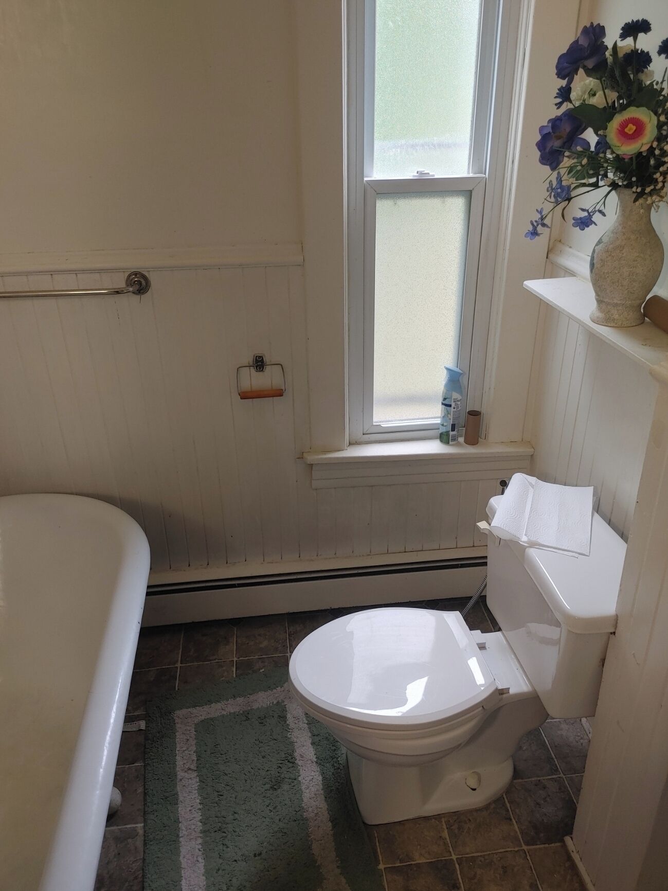 Un baño con inodoro, bañera, ventana y estante con un jarrón de flores.
