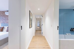 A hotel corridor with a bed on the left, a bathroom with blue tiles on the right, and a framed picture on the wall.