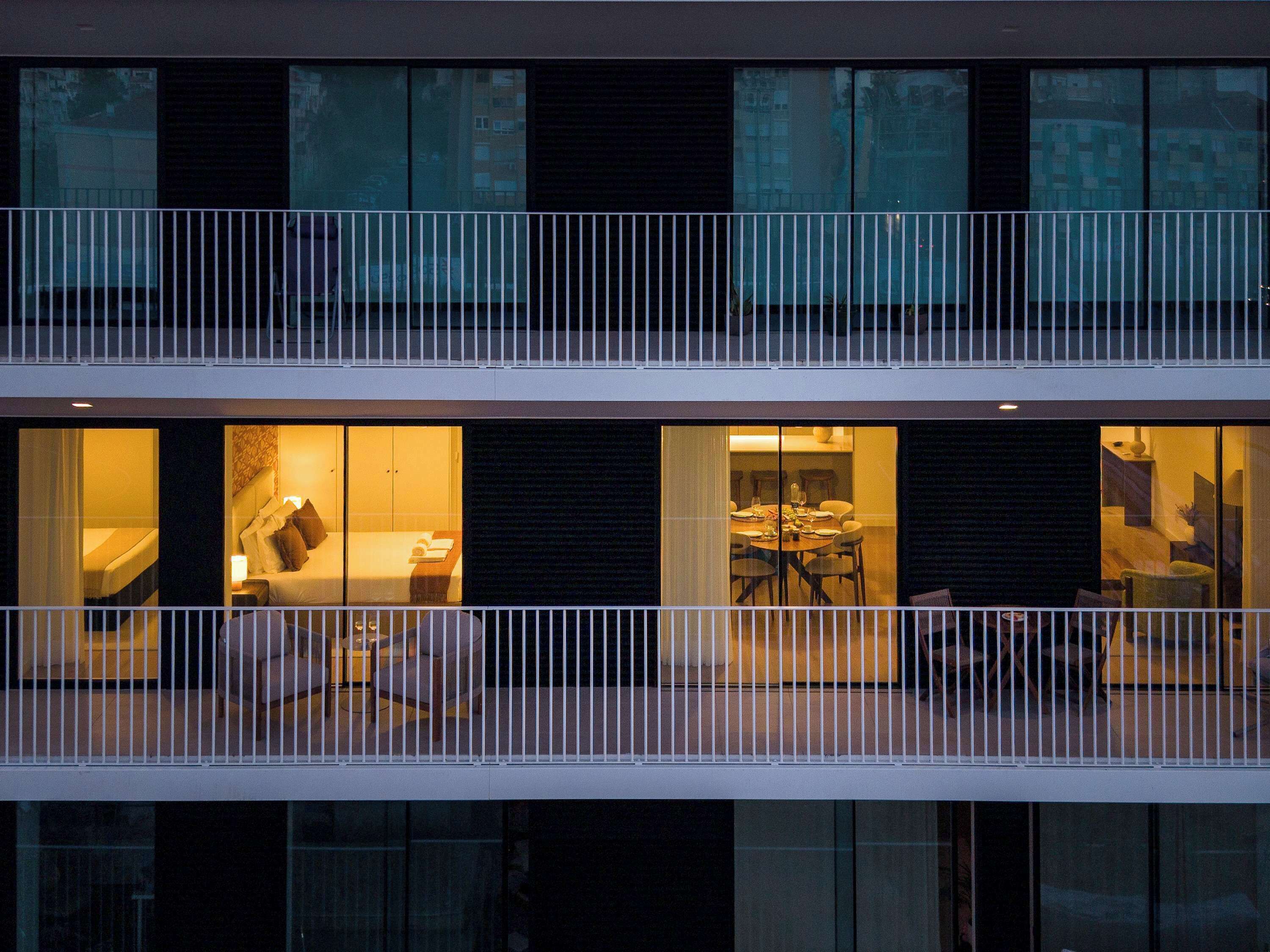 A modern building with a balcony, large windows, and a dining area visible through the glass.